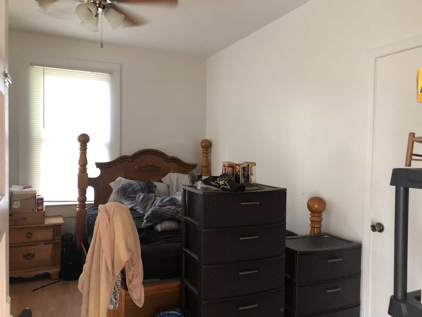 A bedroom with a wooden bed frame, a ceiling fan, two black drawer units, and items scattered on the furniture and floor.