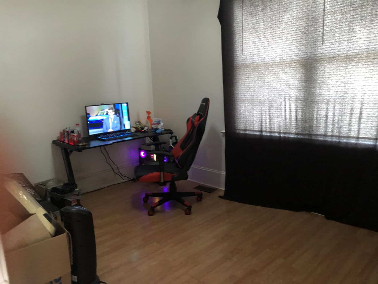 A home office setup with a computer desk, a monitor, various desk items, a red and black gaming chair, and a window covered with a translucent curtain.