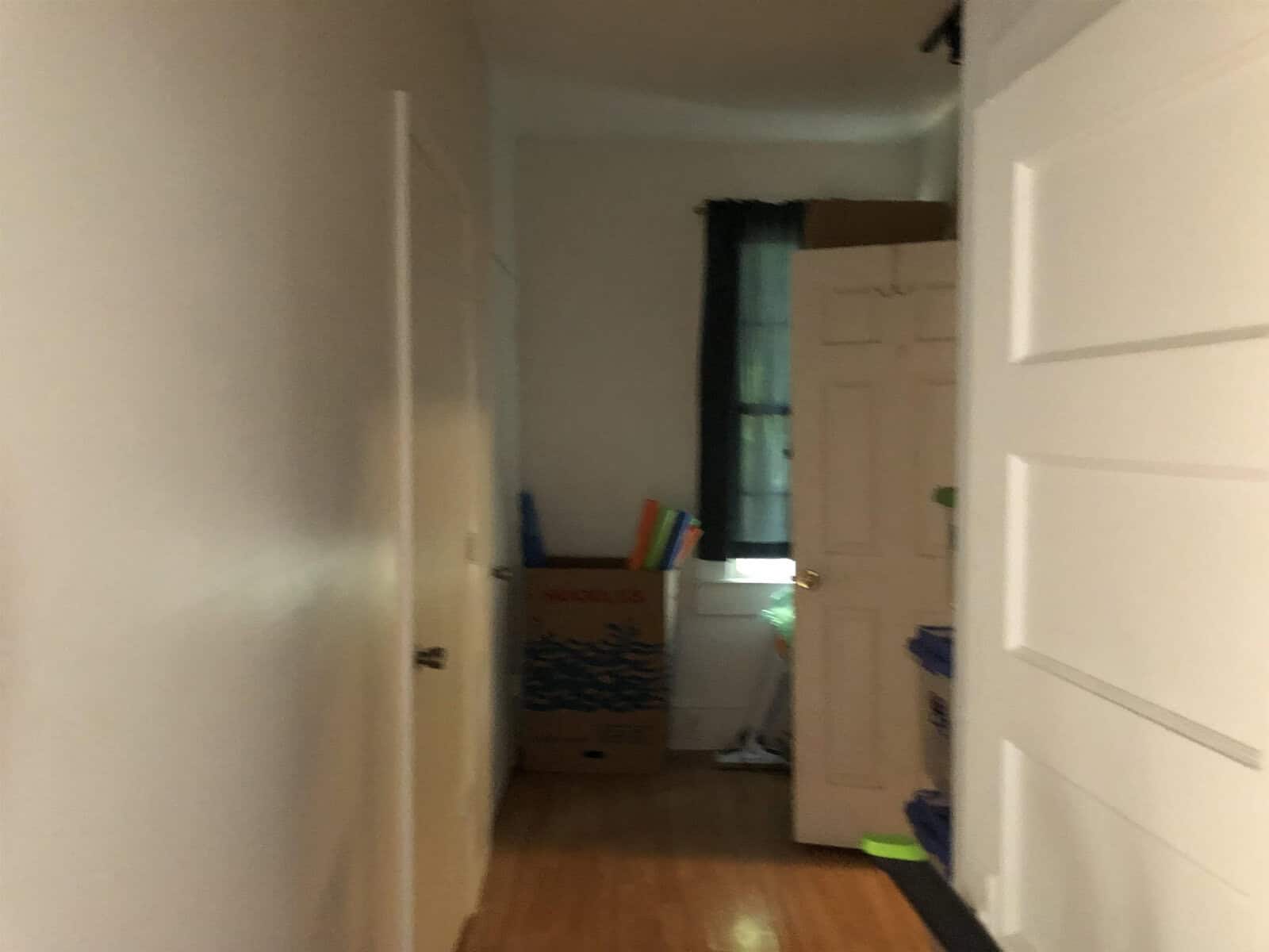 A dimly lit interior hallway leading to a room with an open door, featuring cardboard boxes and visible items suggesting moving or storage.