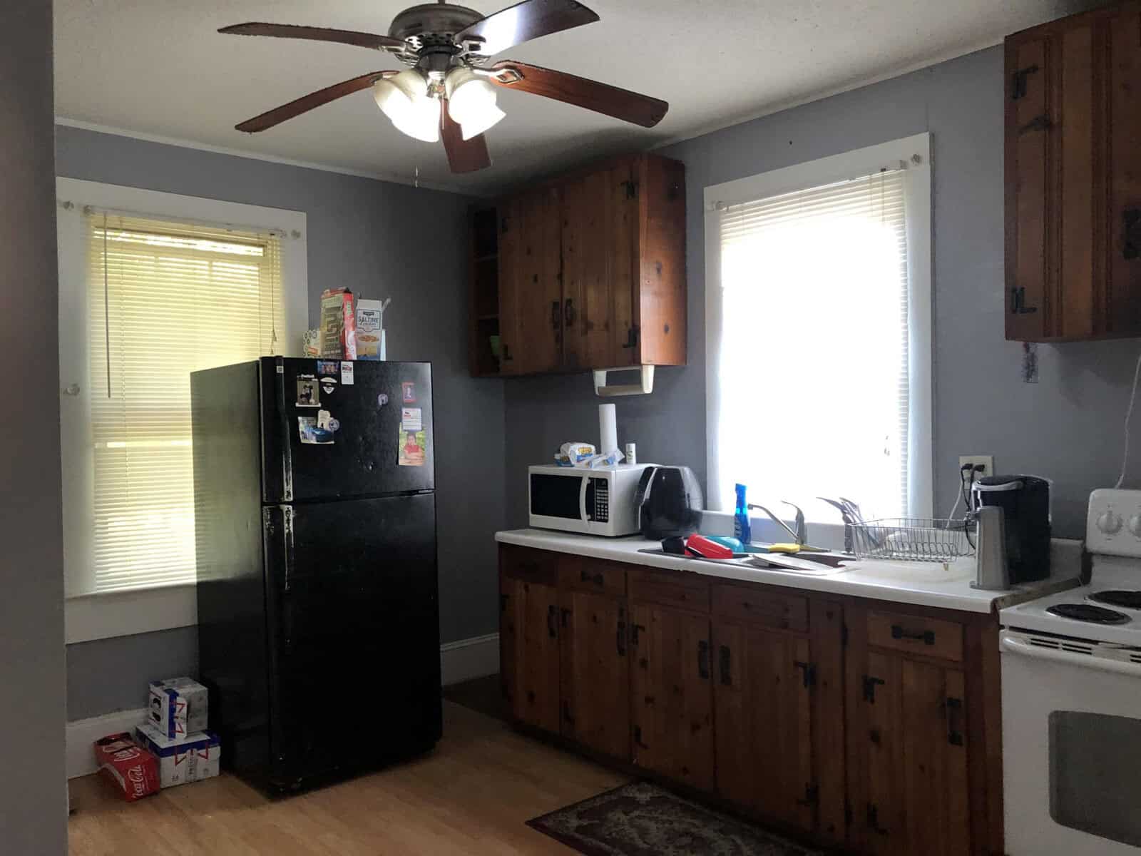 A modest kitchen with wooden cabinetry, black refrigerator, white stove, microwave, and a ceiling fan, illuminated by natural light from a window.