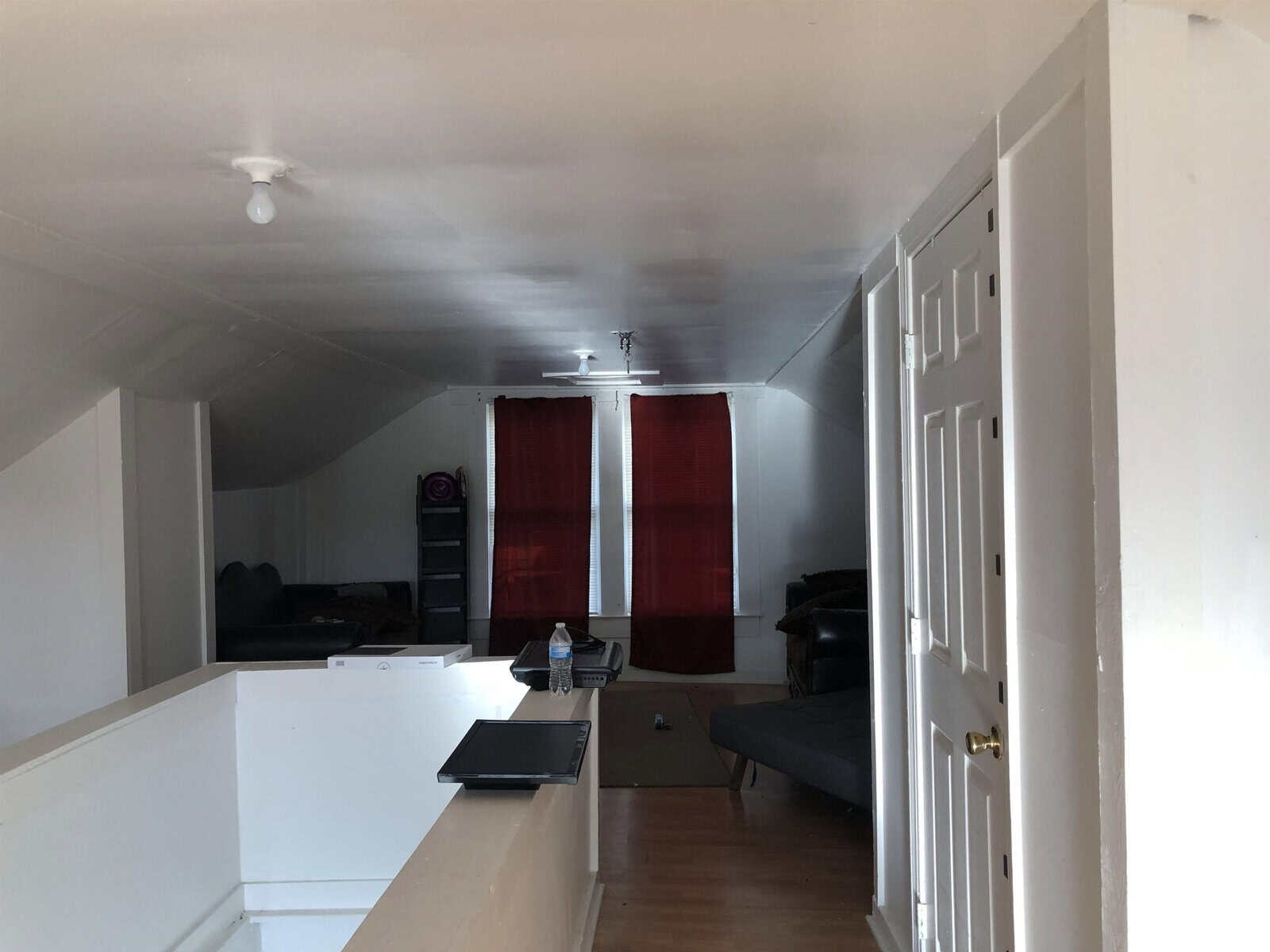 A narrow, elongated room viewed from an elevated position, featuring white walls, a slanted ceiling, and a hardwood floor. a sofa, a closed door, red curtains, and electronic equipment are visible at the far end of the room.