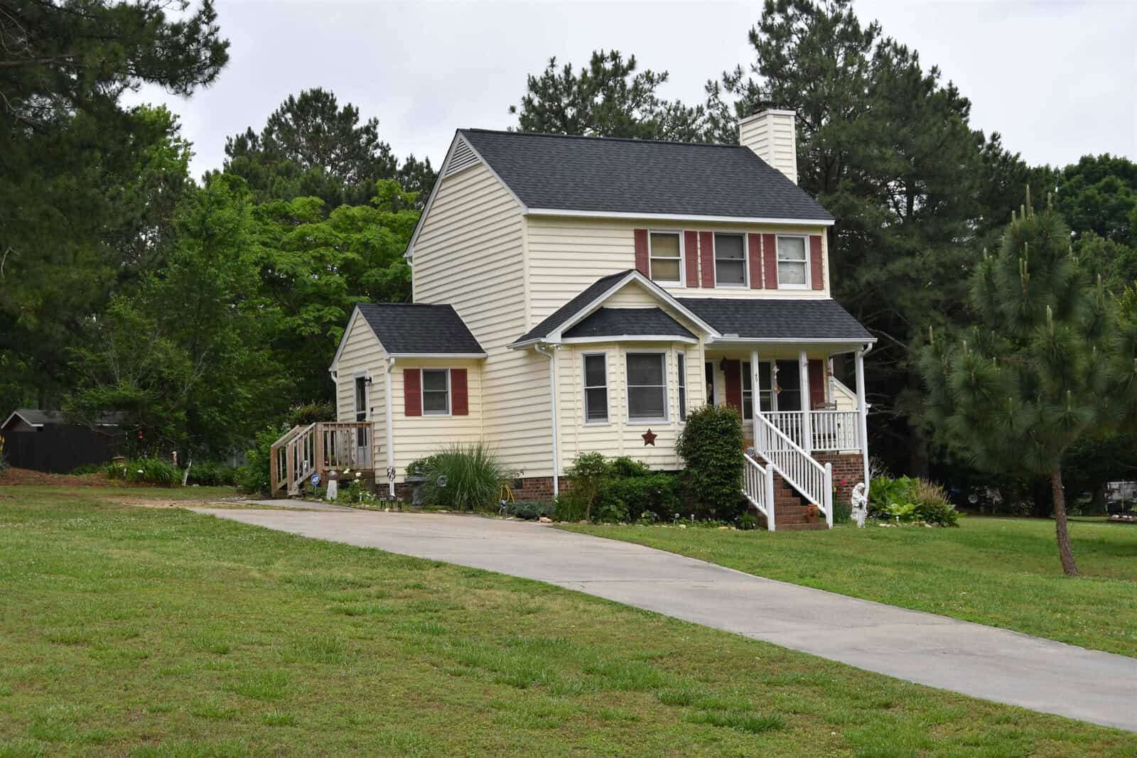 A two-story suburban house with a beige exterior, white trim, and a red door, featuring a front lawn and a paved walkway, surrounded by trees.
