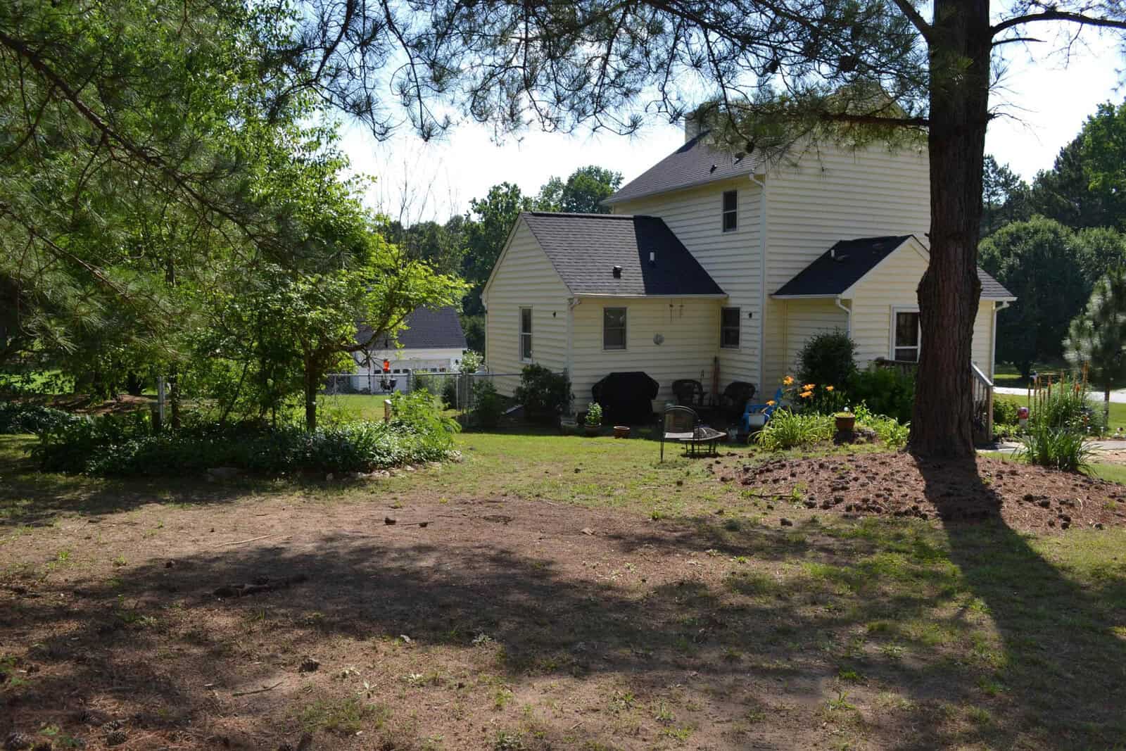 A suburban backyard with a well-maintained lawn, featuring a yellow house with white trim, shaded by trees and equipped with a barbecue grill and outdoor seating.