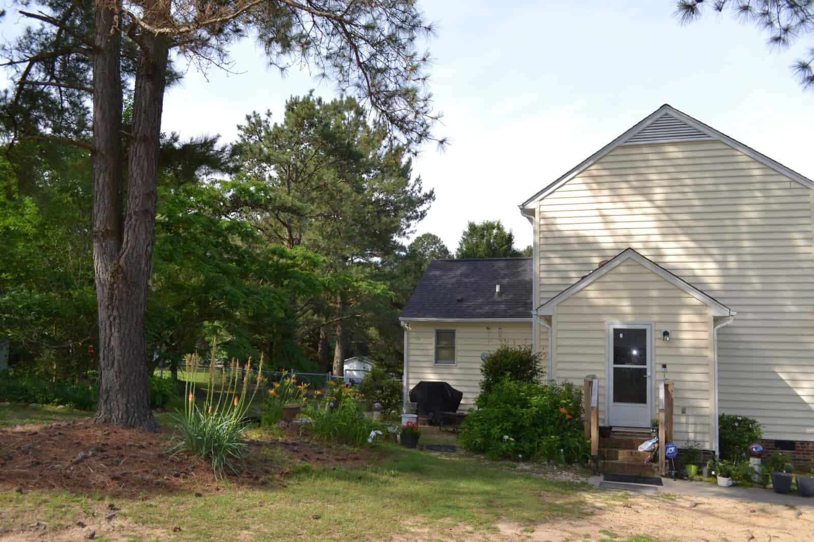 A two-story suburban home with beige siding and a sloped roof, featuring a backyard with a grill, a small garden, and trees under a clear sky.