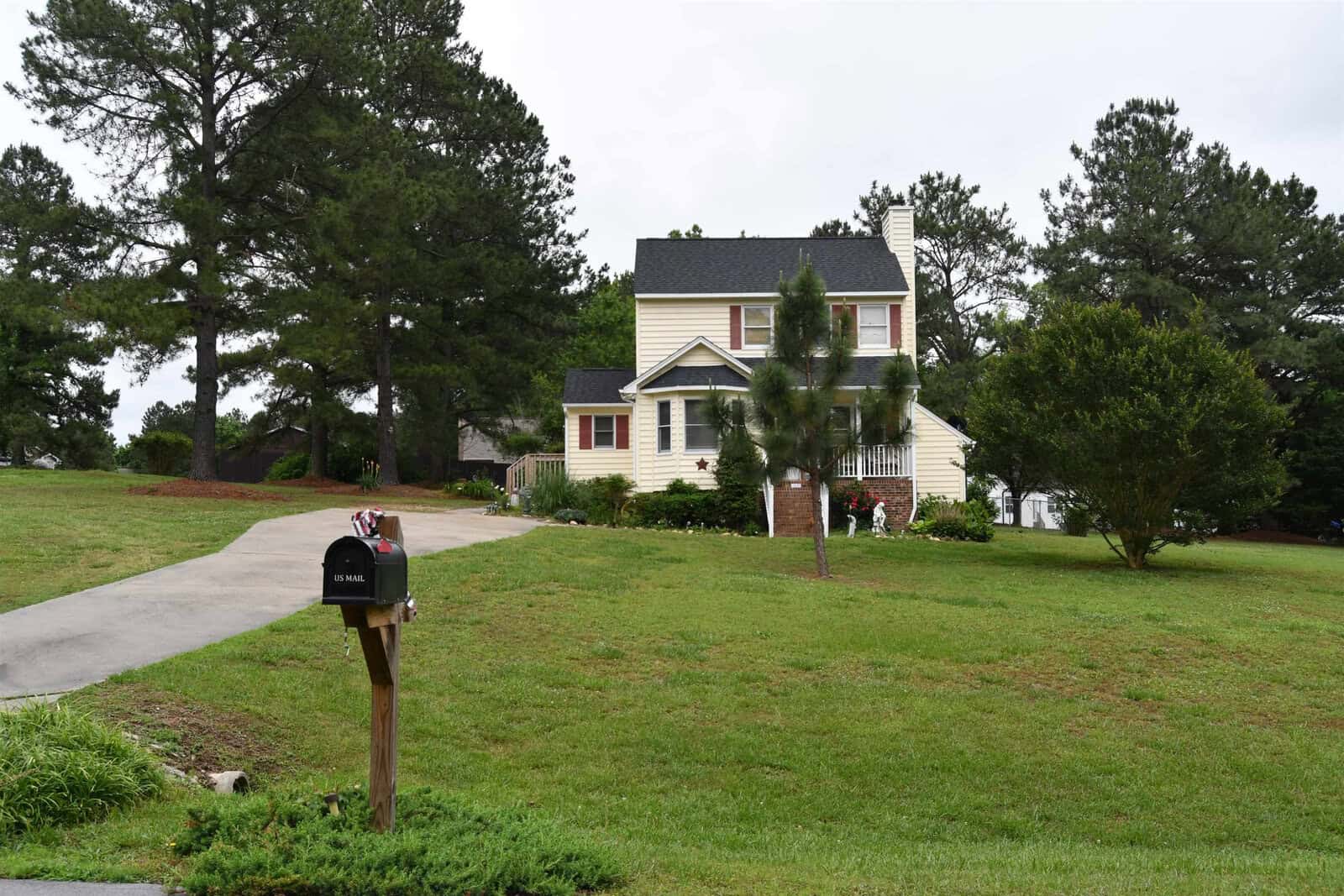 A two-story suburban house with white siding, green shutters, and a gable roof is visible, surrounded by a well-manicured lawn and mature trees. a concrete pathway leads to the entrance, and a mailbox is situated at the foreground on the right side.
