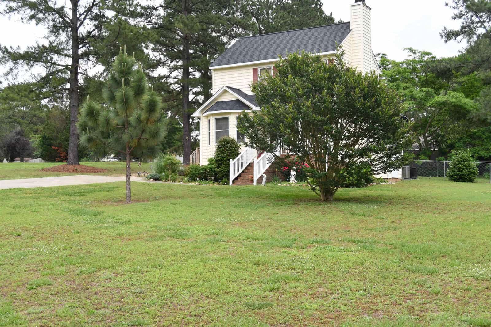 A two-story house with white siding and a brick chimney, featuring a small front porch with white stairs, surrounded by green lawn, bushes, and a couple of trees.