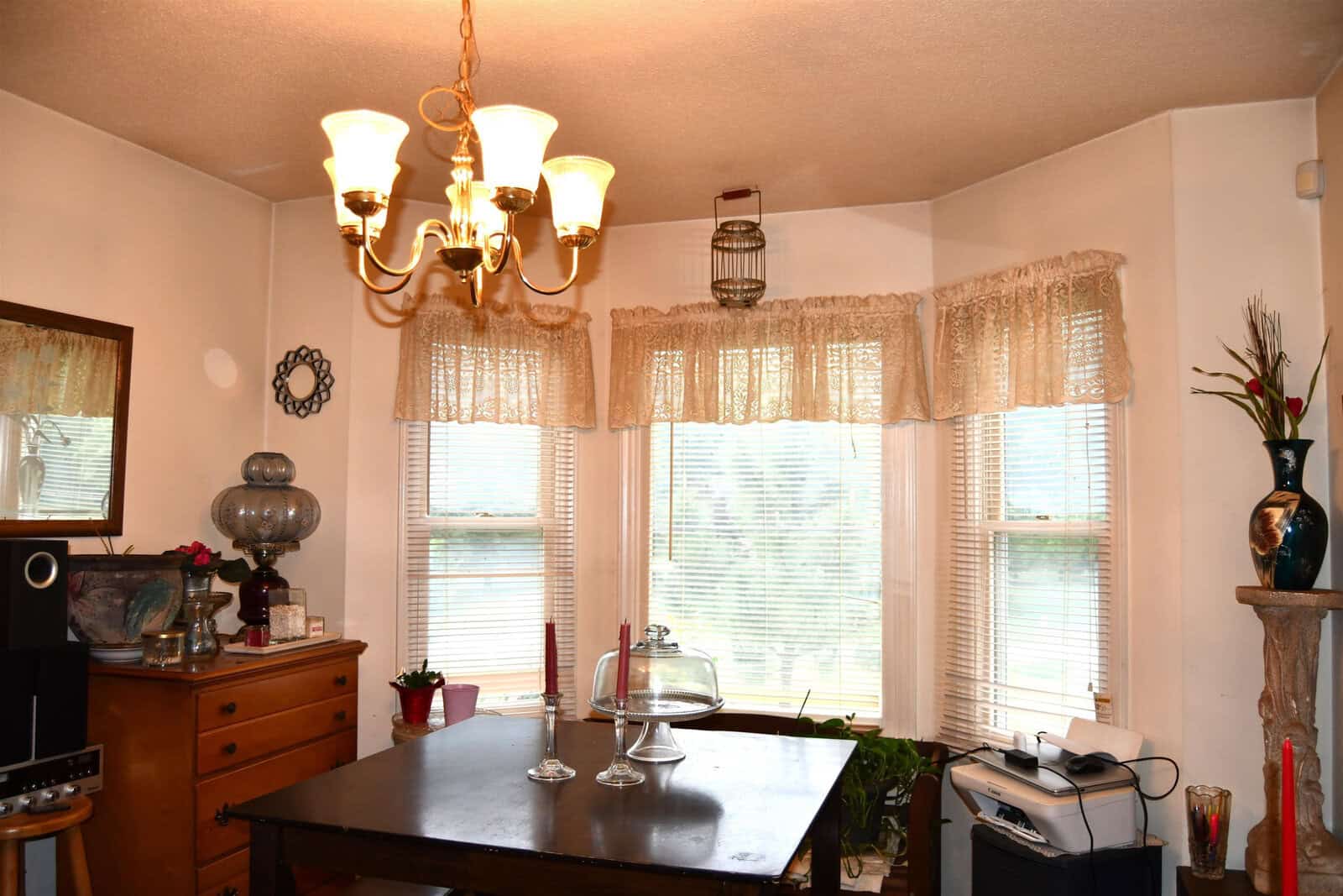 A well-lit dining room with a chandelier, a dark wooden table, sheer curtains on the windows, and various decorative items including a vase and a glass cake stand.