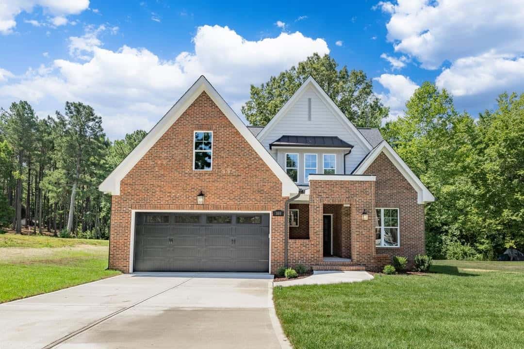 A two-story suburban home at 305 Par Drive, Henderson NC, with a brick facade, featuring a two-car garage and a landscaped lawn under a clear blue sky.