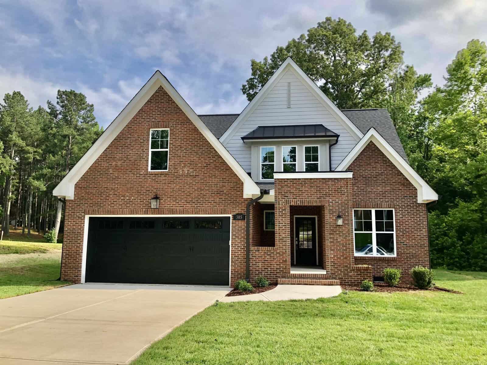 A two-story brick house with a double garage, white trim, and a gabled roof, surrounded by a lawn with trees in the background.