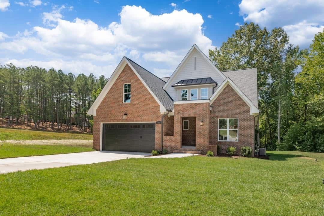 A single-family brick house with a double garage under a clear sky, surrounded by a freshly mowed lawn and a backdrop of trees.