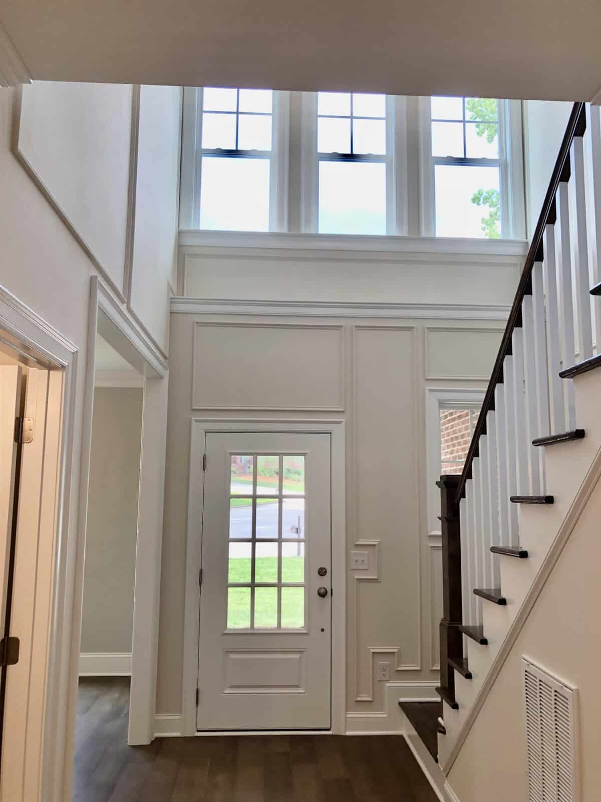 An interior view of a well-lit foyer with a white front door, high windows above the door allowing natural light, a staircase to the right, and wooden flooring.