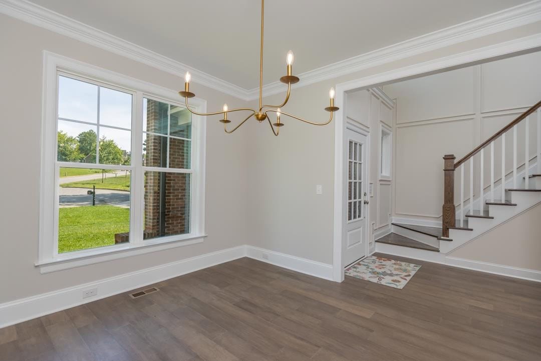 An empty room featuring hardwood floors, a large window with a view of the exterior lawn, a modern chandelier, and a staircase with a wooden handrail leading to the upper level.