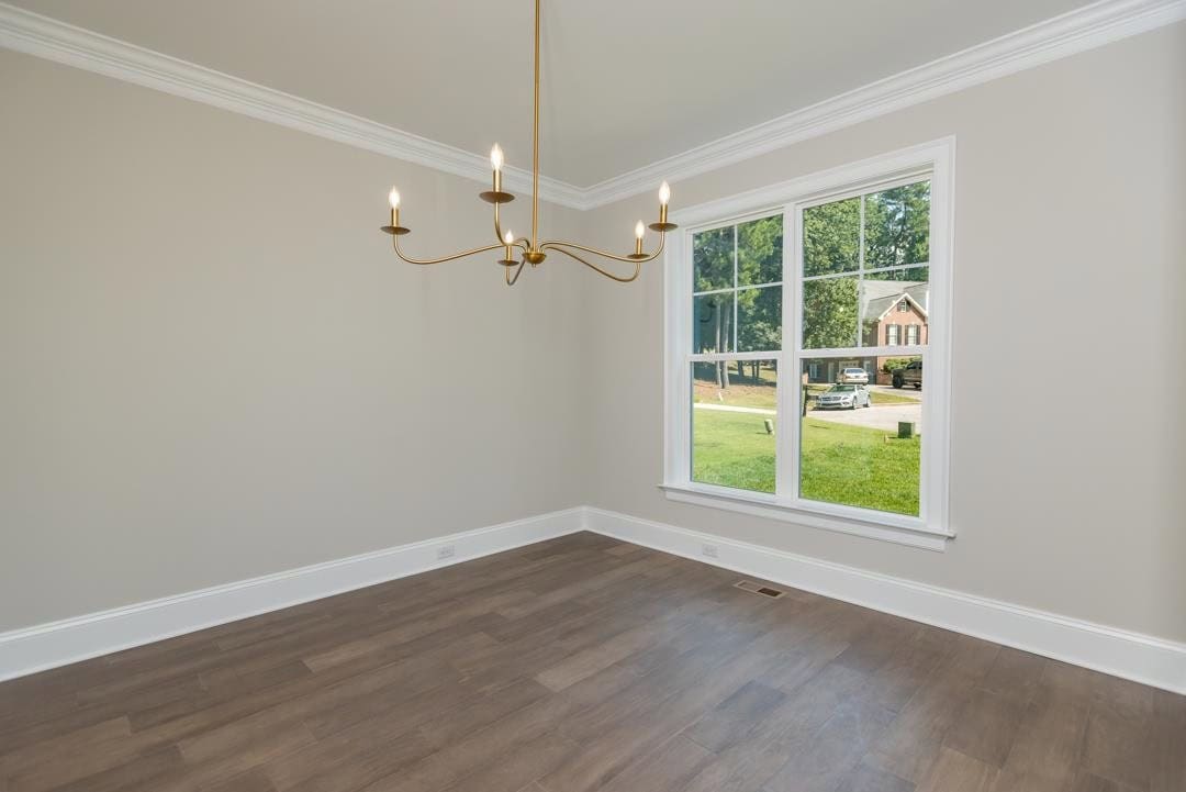 An empty room with beige walls and dark hardwood flooring, featuring a large window providing a view of the outside and a simple chandelier hanging from the ceiling.
