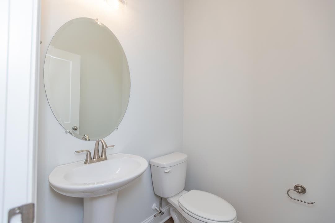 A small bathroom with a round mirror above a pedestal sink, next to a toilet and a towel holder on the wall.