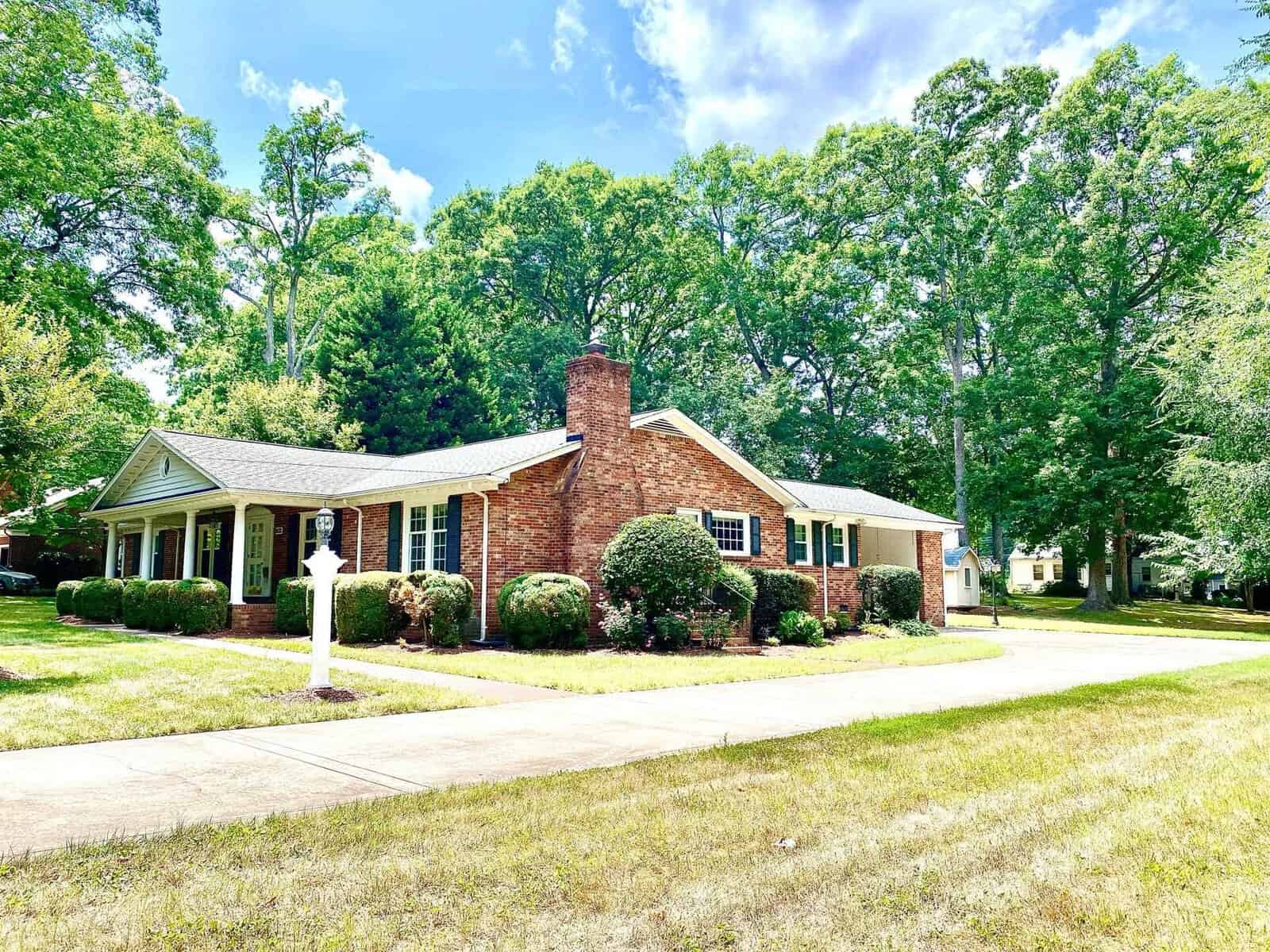 A single-story brick house with white trim, a prominent chimney, surrounded by manicured shrubbery, and a verdant lawn under a blue sky with scattered clouds.