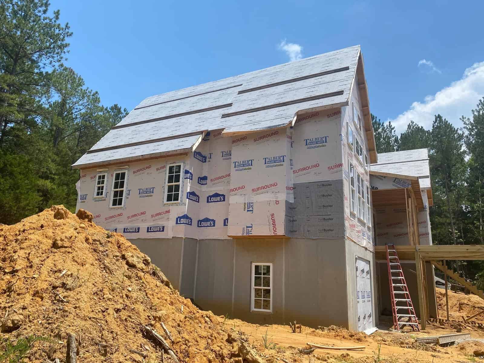 A house under construction with exposed insulation and roofing materials, located in a cleared area with sandy soil and a pile of excavated earth to the side.
