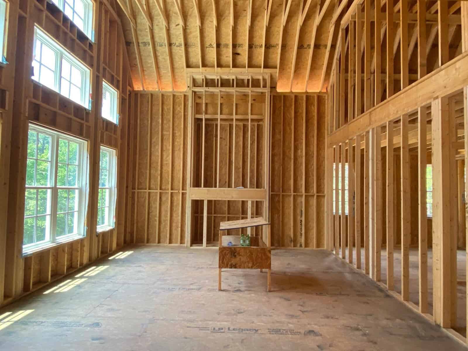 Interior view of an unfinished room with exposed wooden framing and a solitary wooden bench near a window with natural light coming through.
