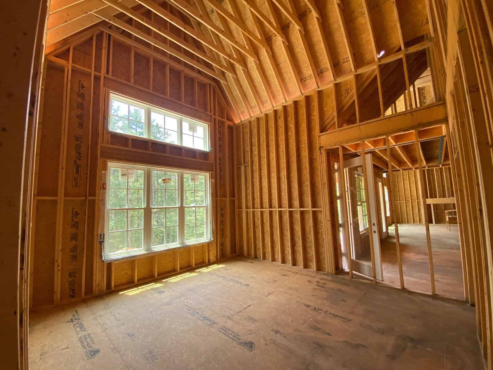 Interior of a house under construction showing exposed wooden studs and framing with a large window letting in natural light.