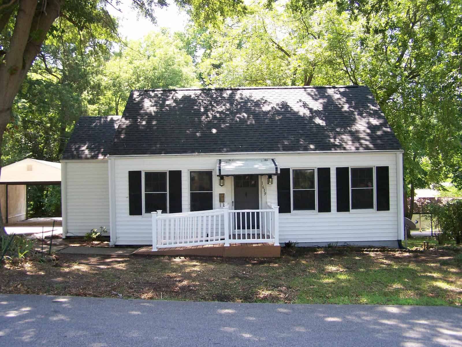 A small white house with black shutters and a dark roof at 1619 Sunset Avenue, Henderson, featuring a ramp to the front door. The home is surrounded by trees and has a driveway with a carport on the left.