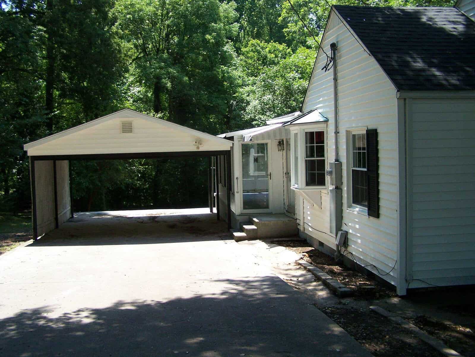 A white house with green shutters and a covered carport sits next to a driveway, surrounded by tall green trees and lush foliage on a sunny day.