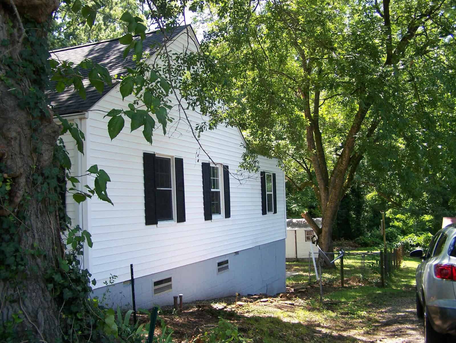 A white house with black shutters sits among green trees on a sunny day. A driveway and part of a parked car are visible on the right, with a small shed in the background.