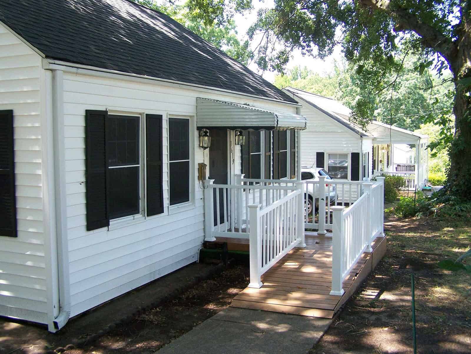 A small white house with black shutters, a covered front porch with white railings, and a wooden ramp. The house is surrounded by trees and is connected to similar neighboring houses.