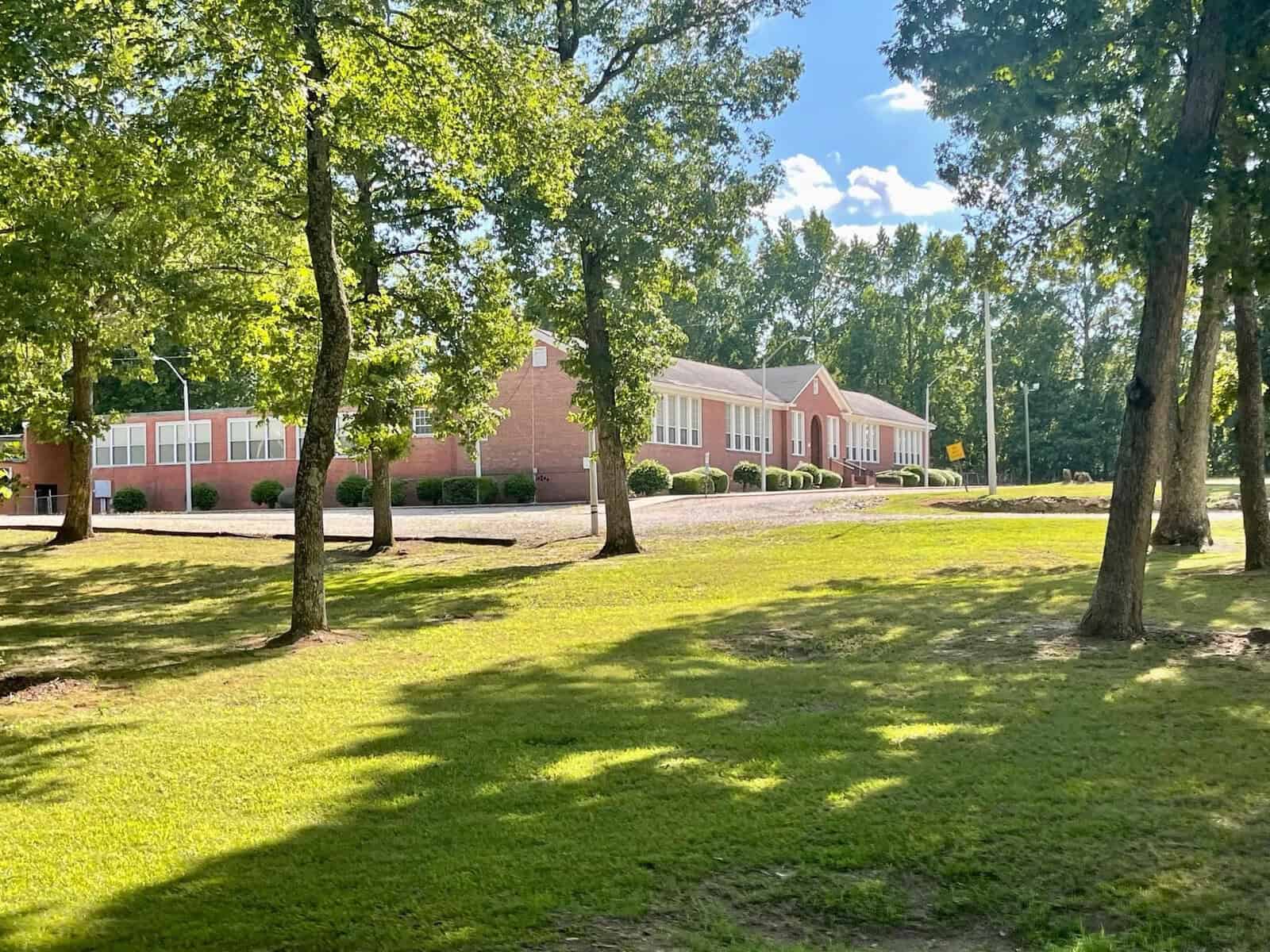 A red brick building with white trim windows, situated behind a grassy area with scattered trees and shadows on a sunny day.