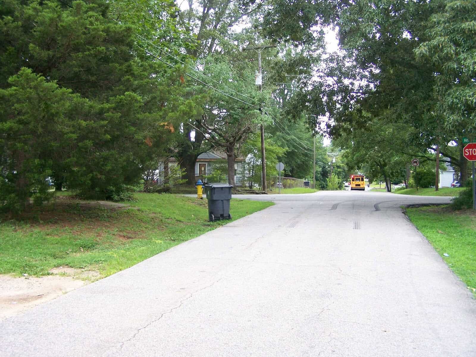 A residential street with a trash bin on the curb, trees lining the road, and a yellow school bus in the distance approaching a stop sign.