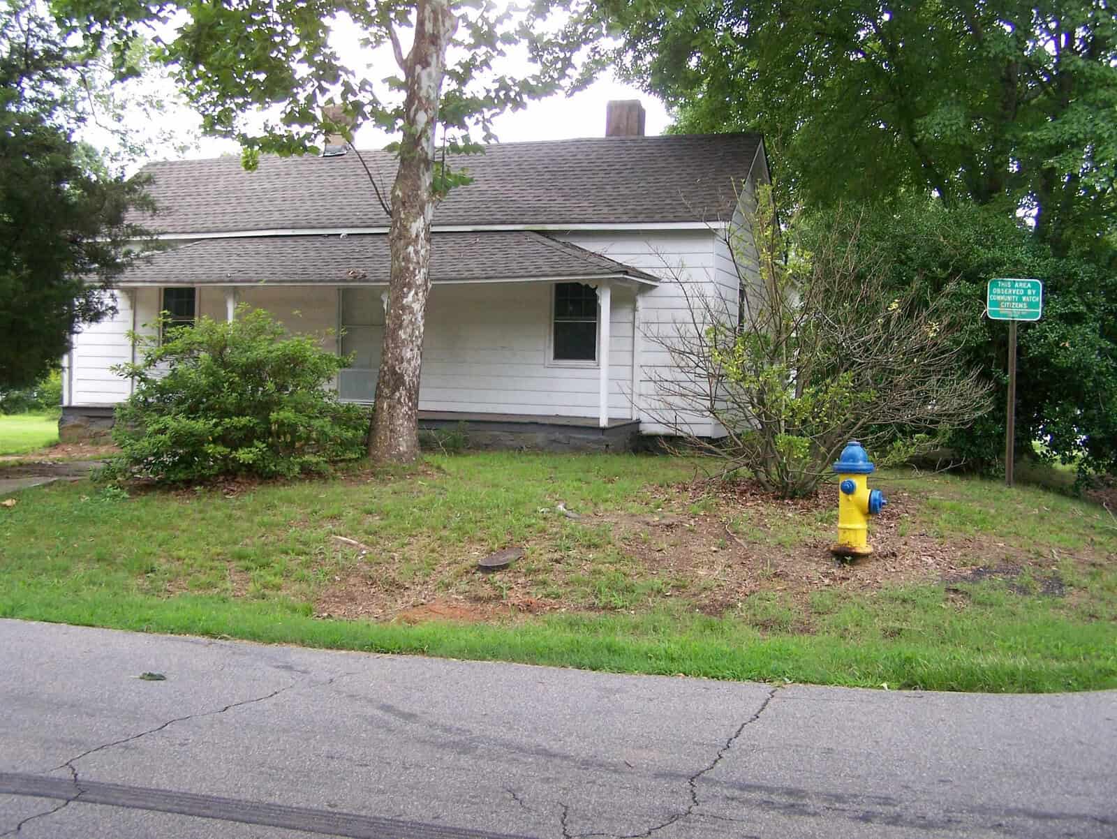 A single-story white house with a grey shingle roof located next to a street, with a yellow and blue fire hydrant in the foreground and a green street sign to the right amidst greenery.