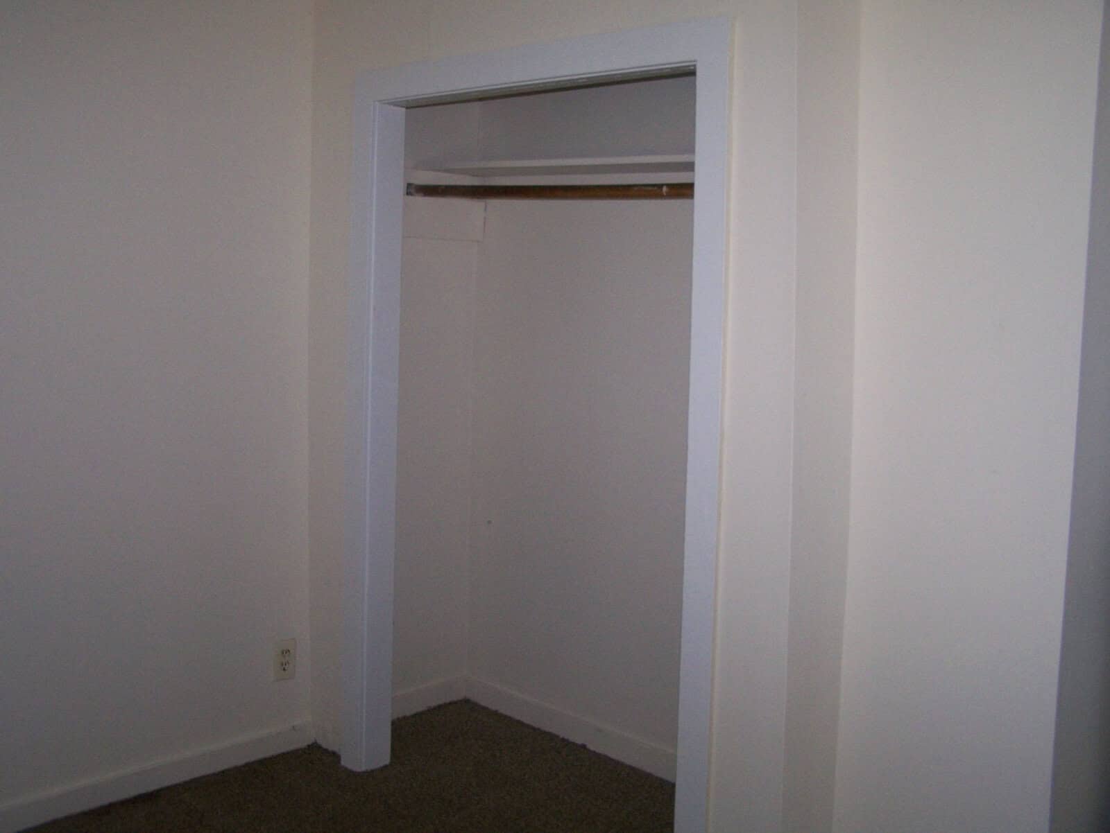 An empty closet with an open door, featuring a single shelf and a hanging rod, set against white walls with a carpeted floor.