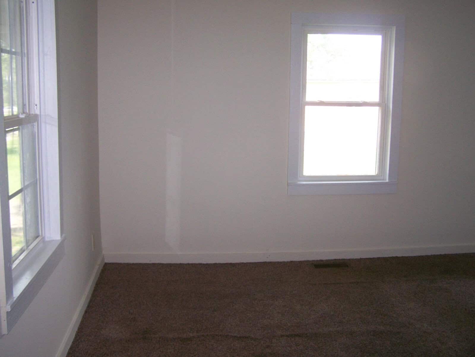 An empty room with a single window and beige carpeting.
