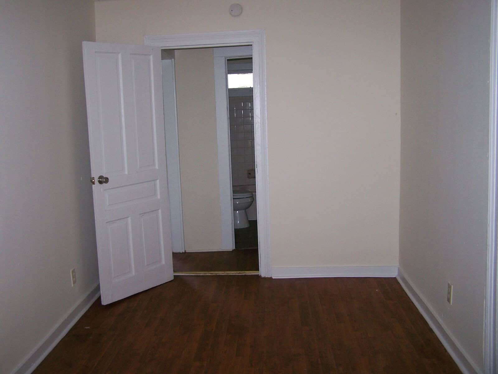 An empty room with wooden flooring, white walls, and an open door leading to a small bathroom with a visible toilet.