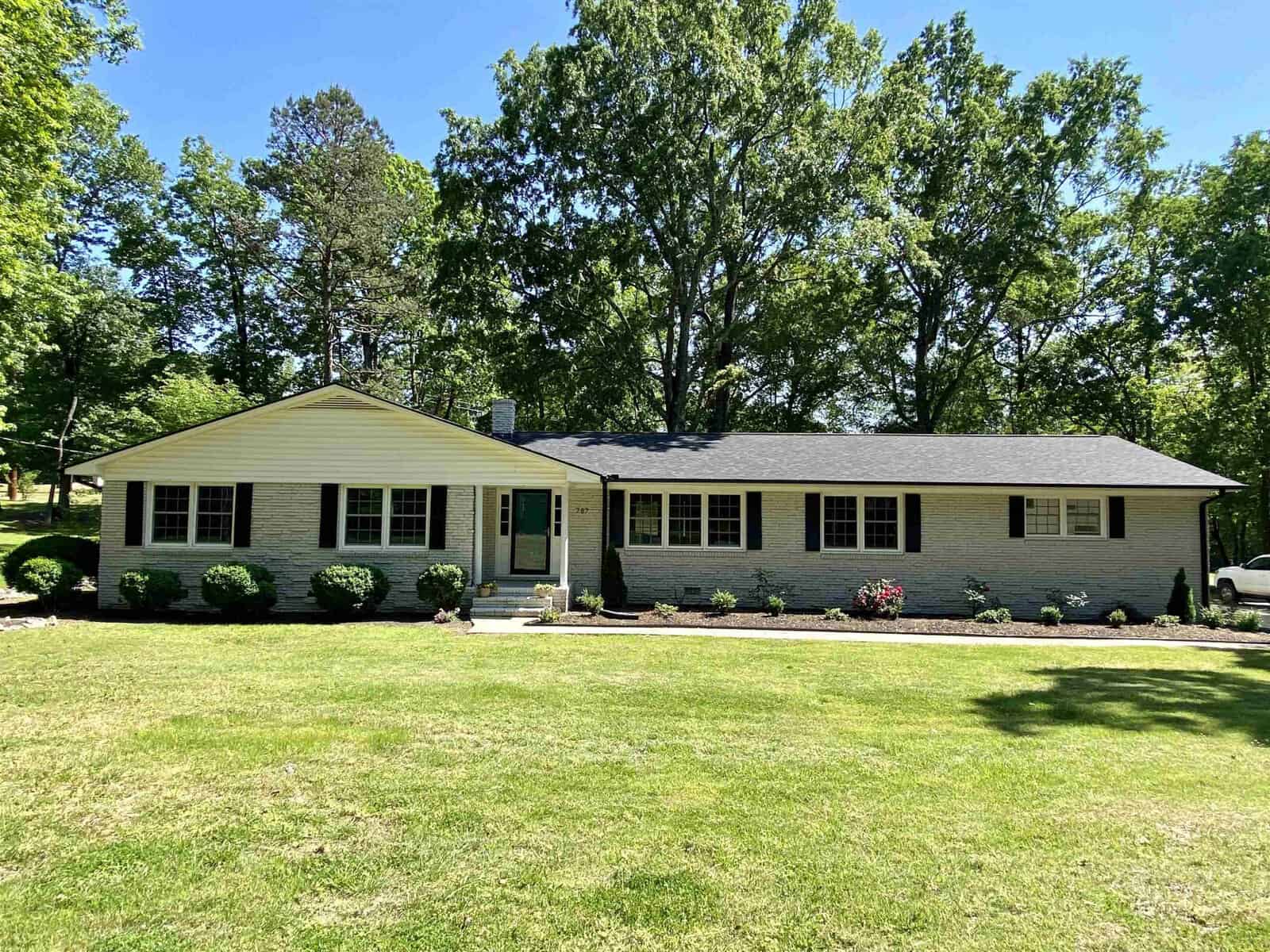 A single-story suburban house at 787 Hedrick Drive, Henderson NC, with beige siding, black shutters, a gray shingled roof, and a well-manicured lawn with a