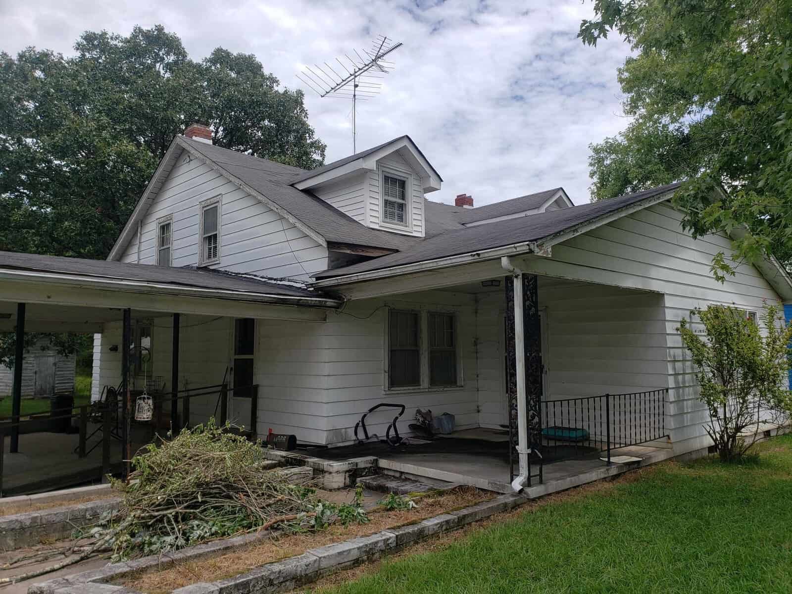 A white, two-story house at 1229 W NC 158 Highway in Oxford boasts a black roof and large TV antenna. It features a covered porch, enveloped by lush greenery and trees, with branches scattered on the lawn. The partly cloudy sky adds to its charm.