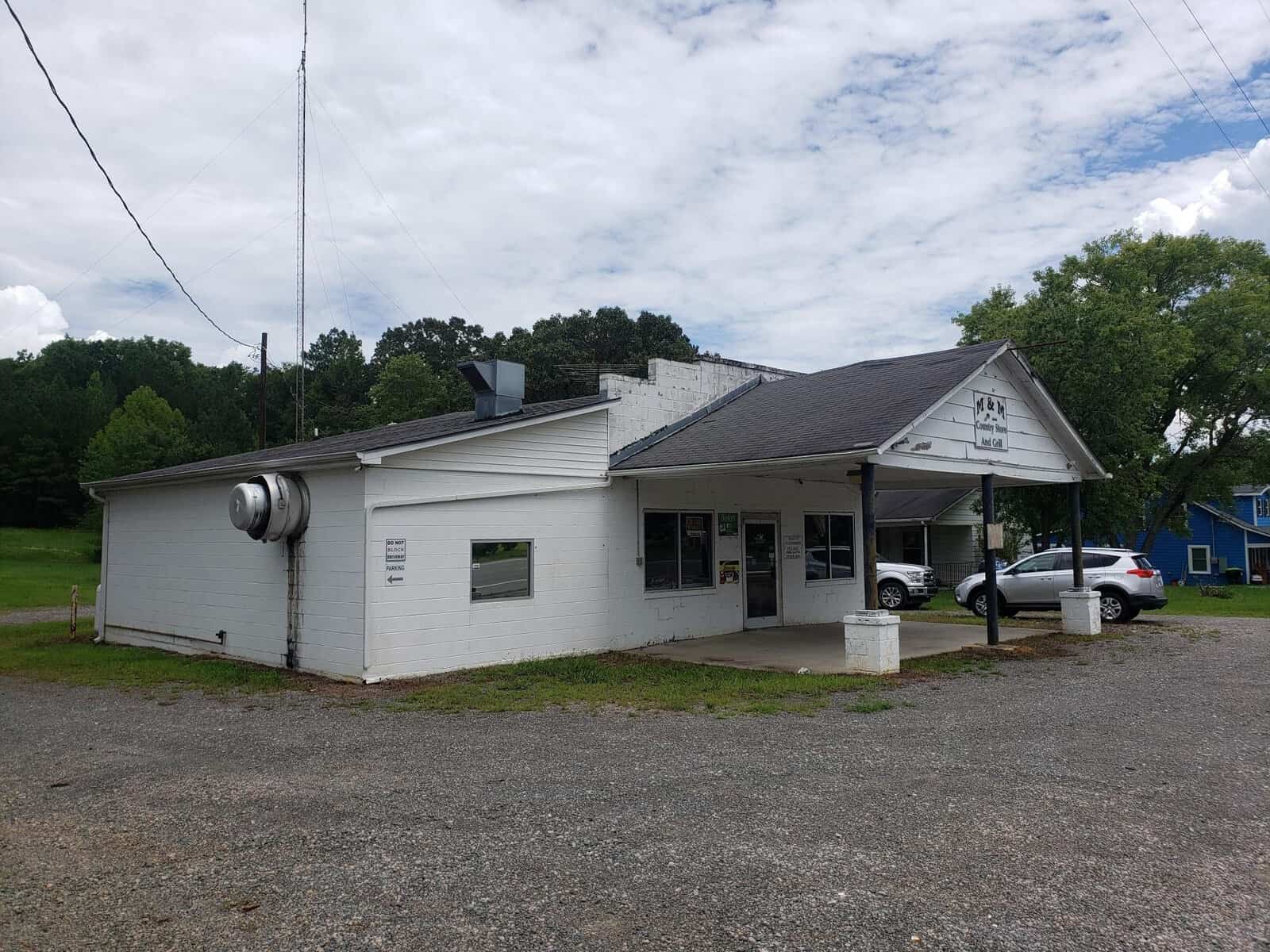 A small, single-story white building with a gabled roof. It has a covered entrance supported by pillars and a vent on the side. The surrounding area is gravel with parked cars and trees in the background under a partly cloudy sky.