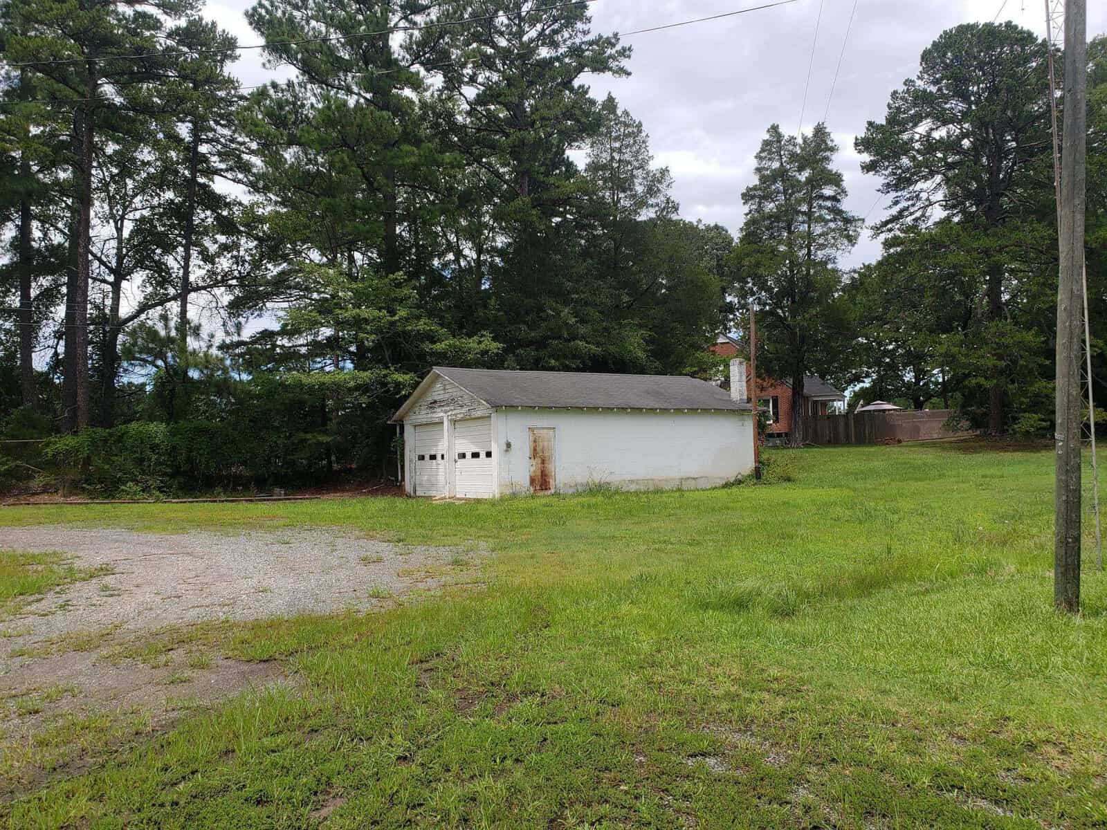 A small, old white garage with two doors stands in a grassy, open area. Tall trees surround the structure, and a brick building is partially visible in the background. Overcast skies give the scene a serene, rural feel.