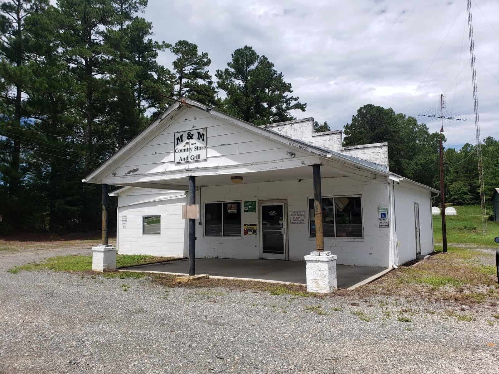 A small, white country store with a rustic appearance, surrounded by trees. The sign above the entrance reads "M & M Country Store and Grill." The gravel parking area is empty, and the sky is overcast. .
