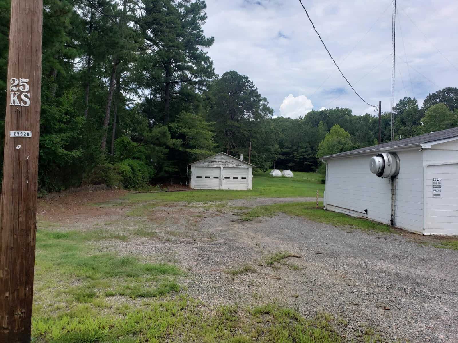 A small white garage with two doors stands among trees and grass. A gravel path leads to the building. To the right, part of a larger white building is visible. A utility pole with markings is in the foreground. The sky is cloudy.