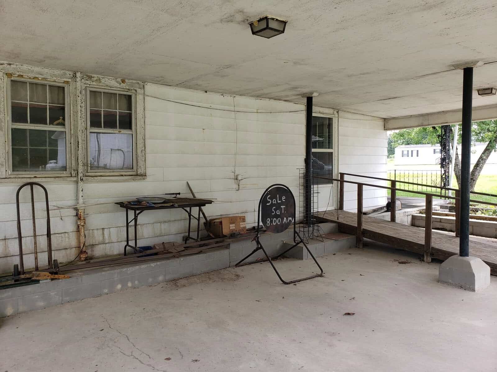 A covered porch with a ramp and a "Sale Sat. 8:00 AM" sign. Old windows and various items like a workbench, tools, and gardening equipment are visible. The area has an outdoor, rustic appearance with a fenced yard in the background.