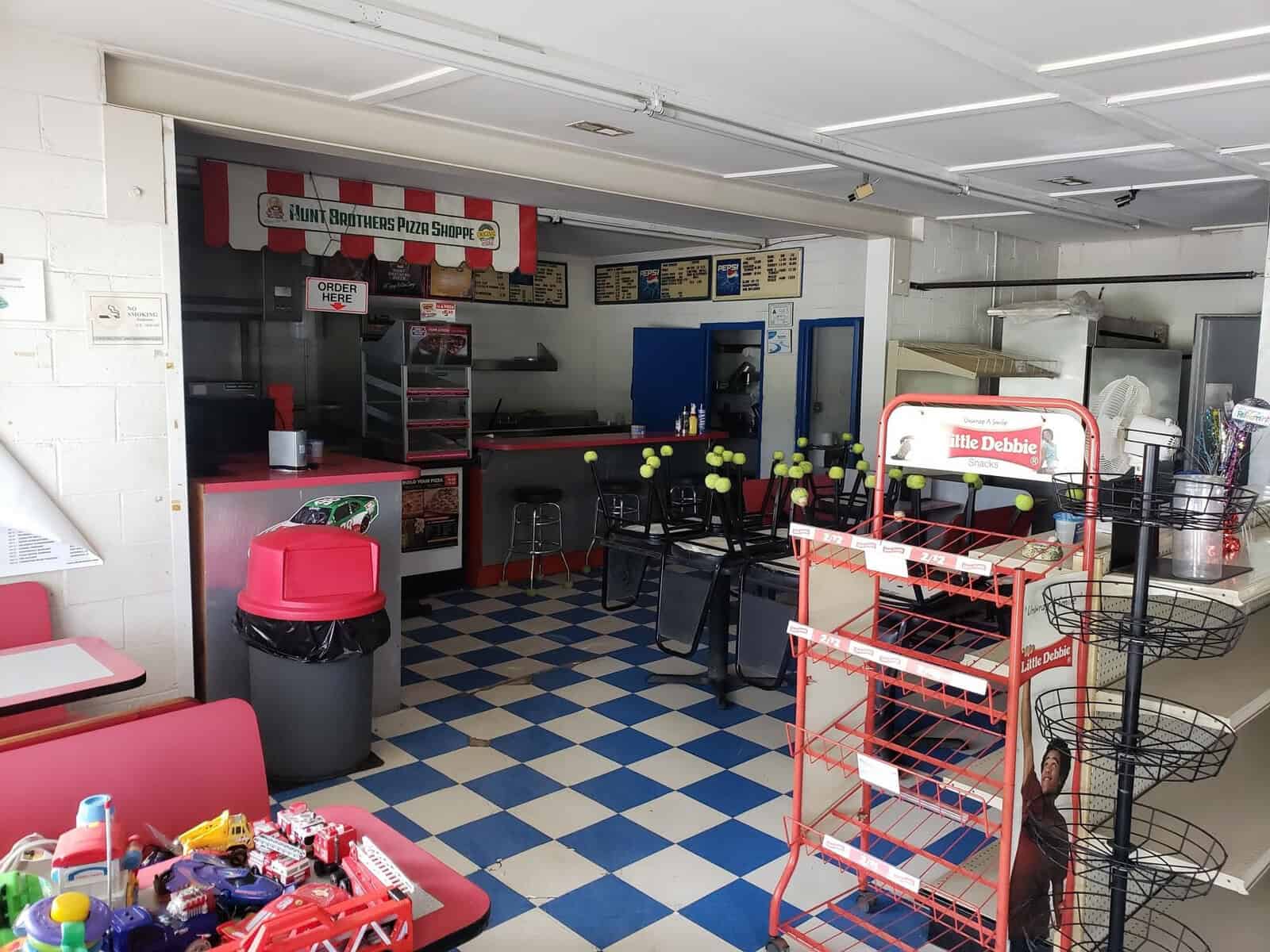 An empty pizzeria with blue and white checkered floors, red booths, and a counter with a "Hunt Brothers Pizza" sign. There's a stand holding empty metal racks and some scattered tables and chairs. Small toys are visible on a nearby table.