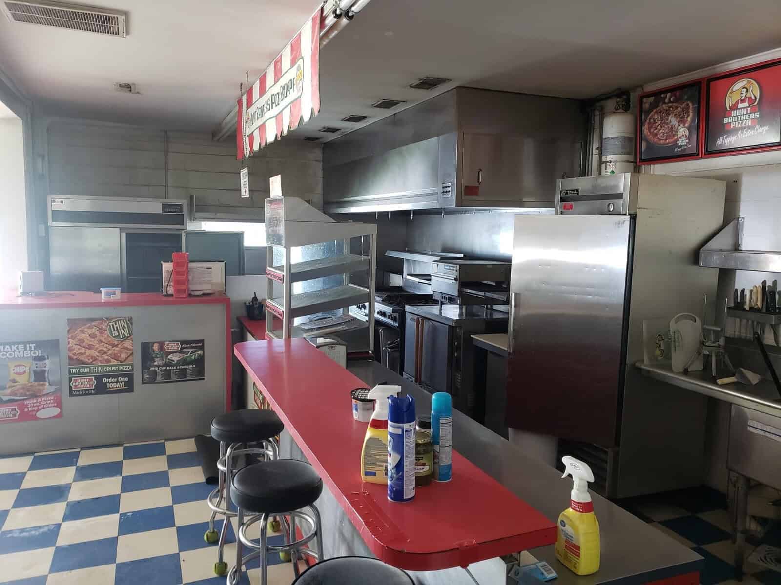 A small, empty fast-food restaurant kitchen with a red counter and metal stools. Cleaning supplies are displayed on the counter. Stainless steel appliances and pizza ovens are visible, along with promotional posters on the walls. The floor is blue and white checkered.
