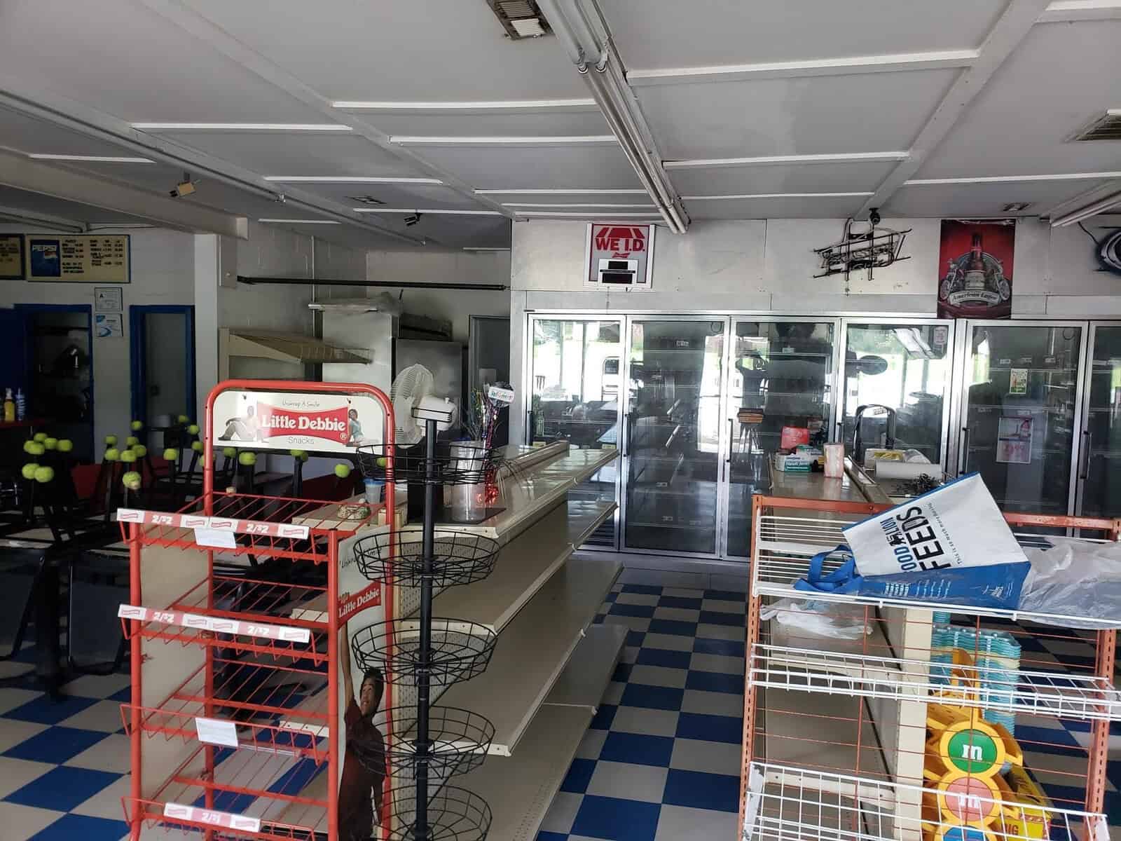 Empty store interior with multiple shelves, a checkered blue-and-white floor, and a wall-mounted cooler at the back. Shelving units are mostly empty, with a few items, including a Little Debbie display rack.
