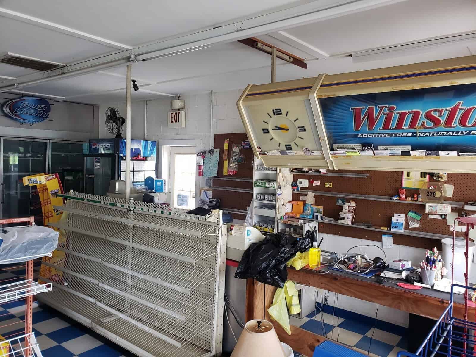 Interior of a convenience store with empty shelves, a clock displaying "Winston," and a check-out counter. Items like pens, a lamp, and cleaning supplies are scattered on the counter. A cooler in the background displays refreshments.