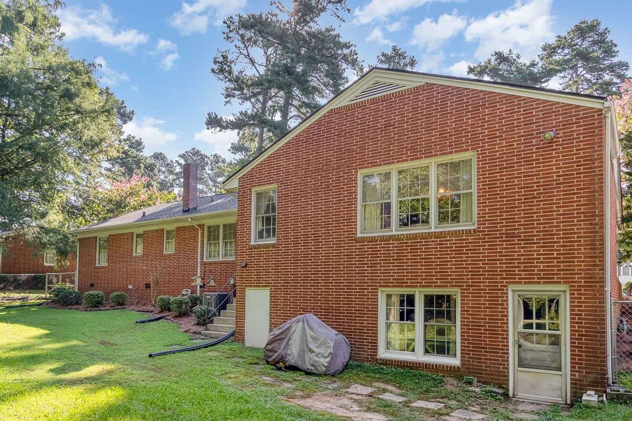 A two-story red brick house with white windows, surrounded by trees and a lawn, under a blue sky with scattered clouds.