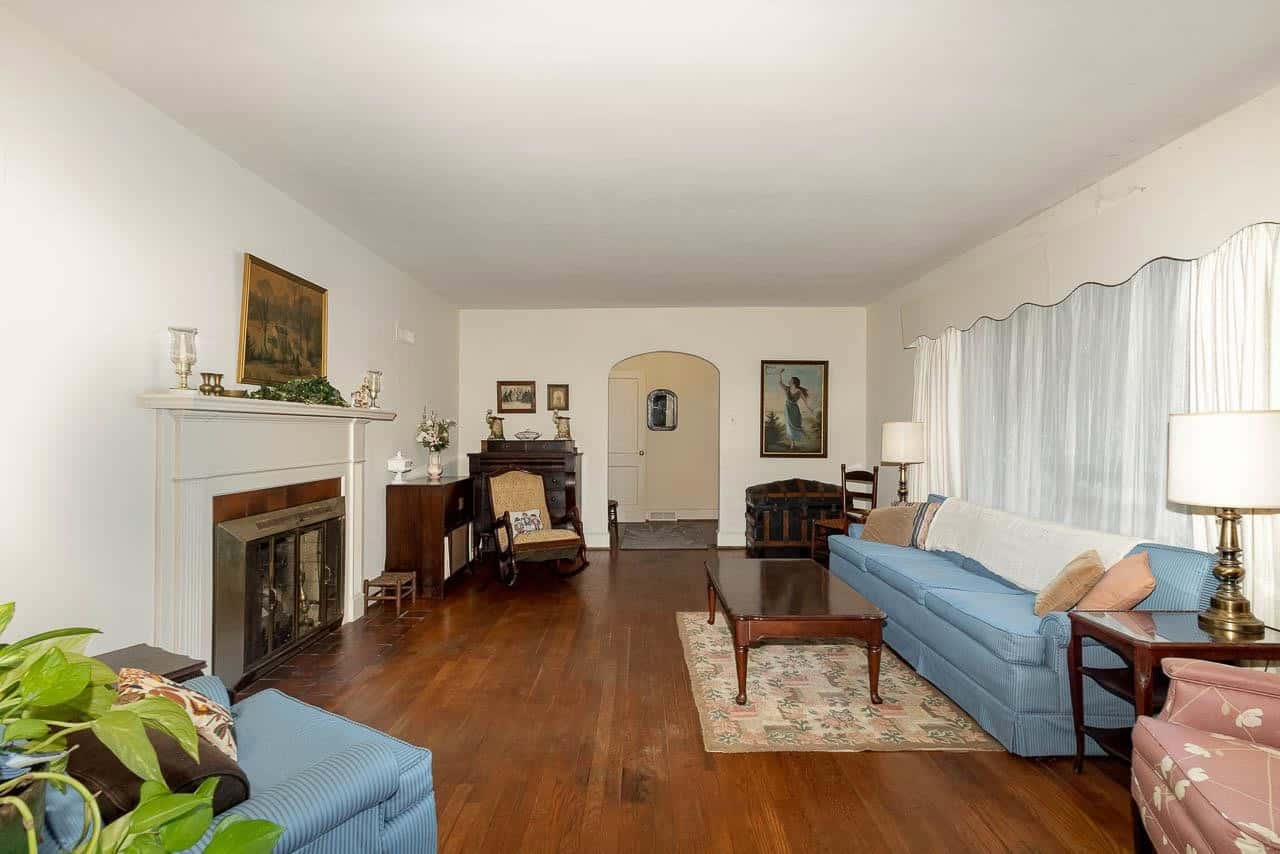 A spacious living room with hardwood floors, featuring two blue sofas, a fireplace, various decorative items on the mantel, and artwork hung on the walls.