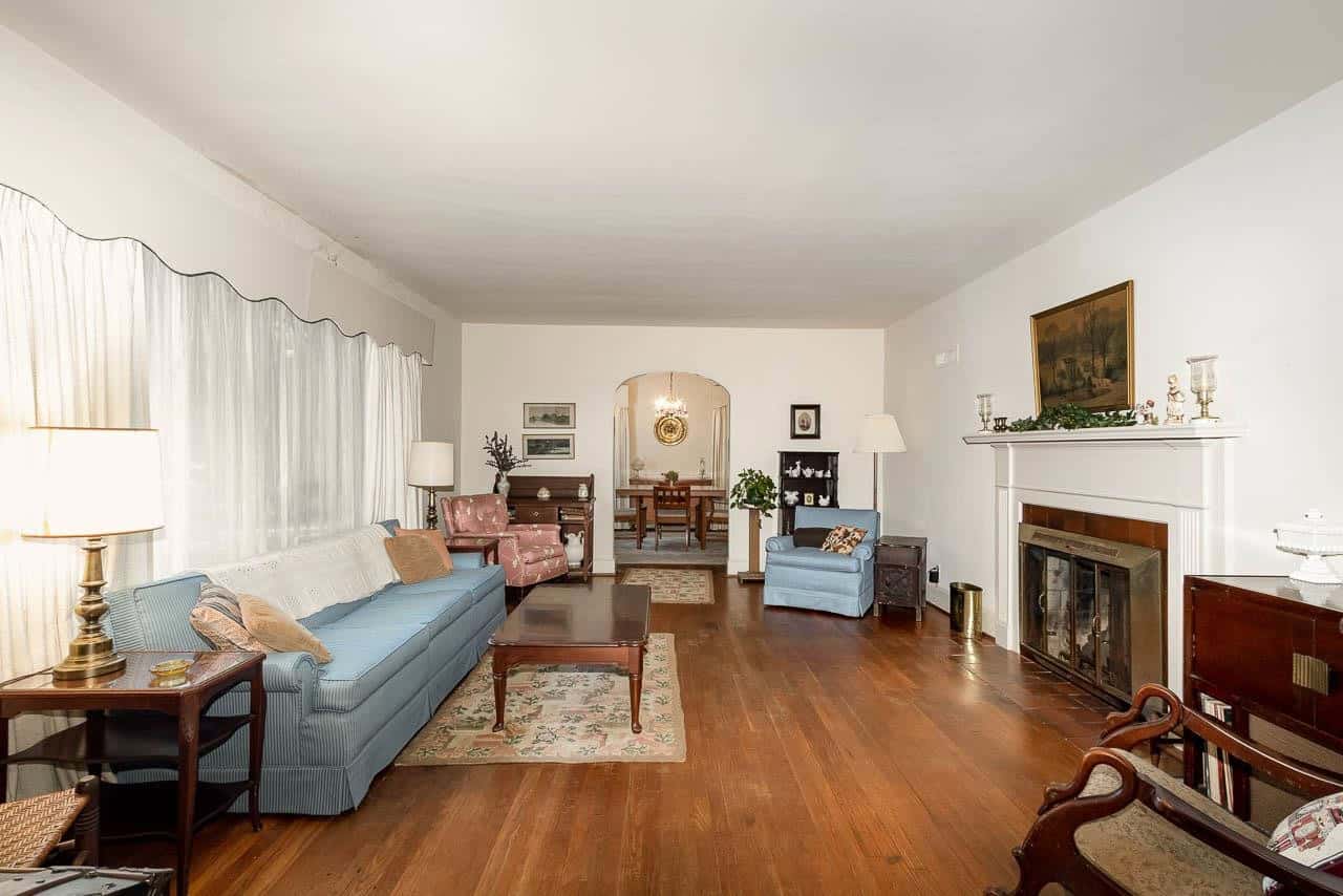 A well-lit, spacious living room featuring hardwood floors, a blue sofa, a wooden rocking chair, a fireplace, and various decorative items arranged neatly throughout the space.