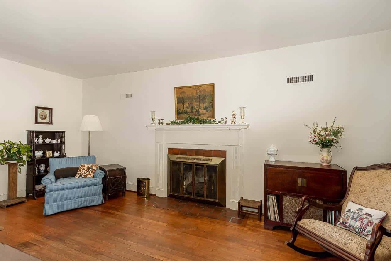 A neatly organized living room with a white fireplace, hardwood floors, and a classic furnishing style, featuring a blue sofa, floral-patterned armchair, and a decorative mantlepiece.