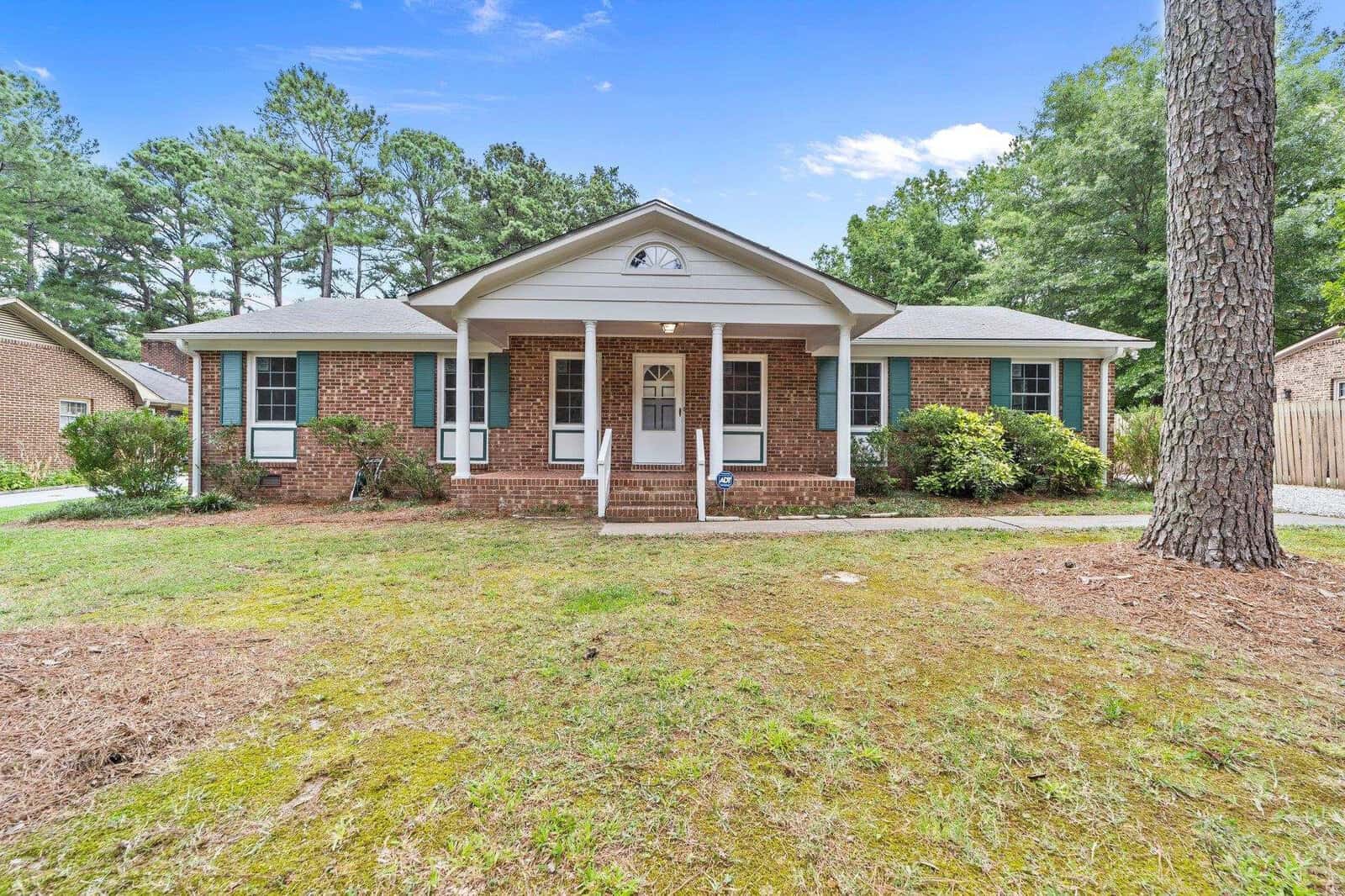 A single-story brick house at 1214 Anne Street, Henderson NC, with a centered front door and symmetrically positioned windows under a gabled roof, surrounded by a lawn with scattered trees.