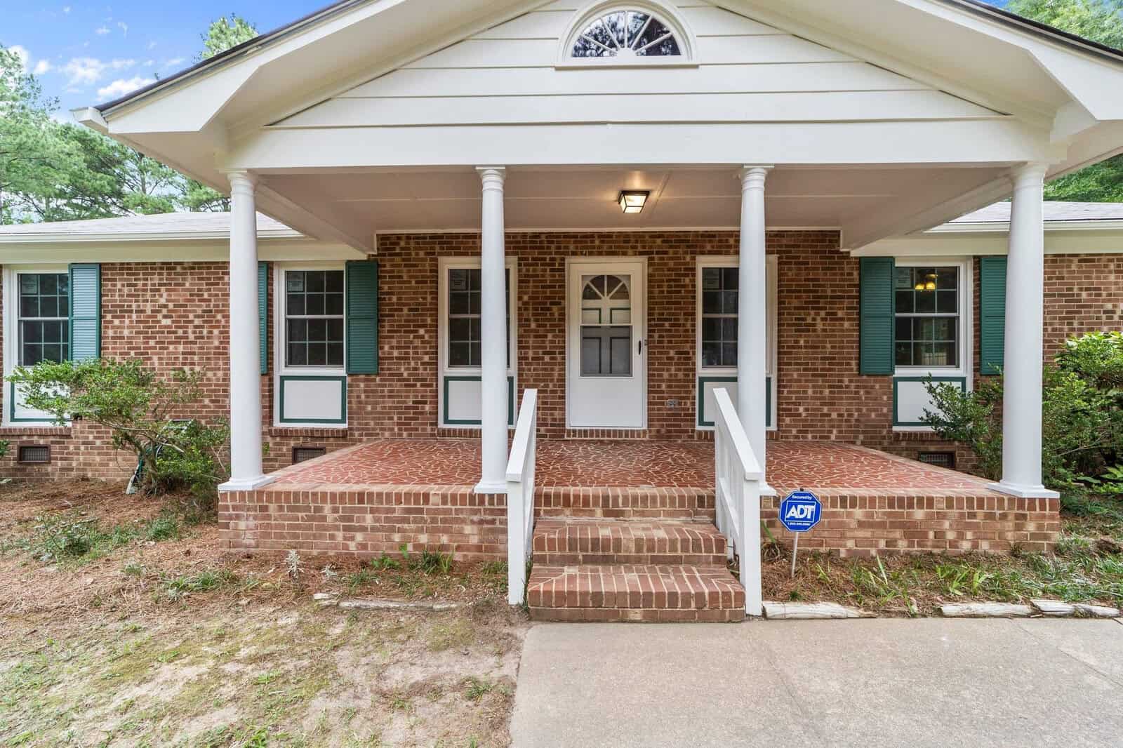 Front view of a single-story brick house with a covered porch, white columns, and an adt security sign on the lawn.