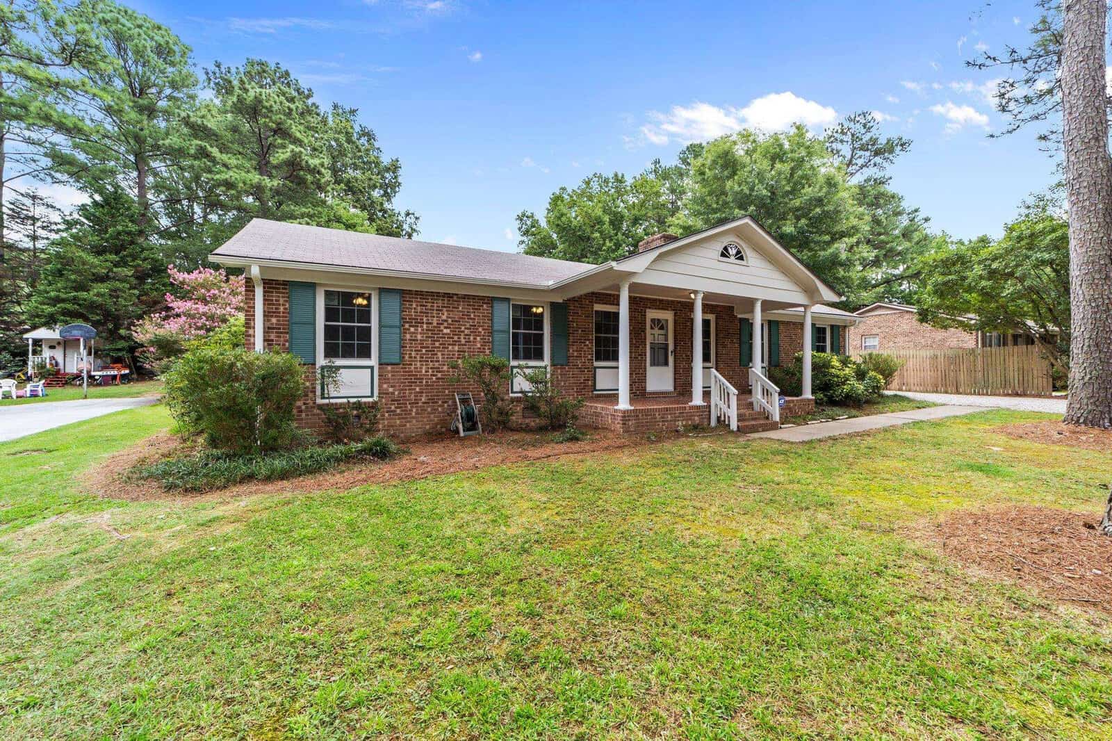 A single-story brick house with white trim, a covered entrance, and a front lawn with trees and shrubs.